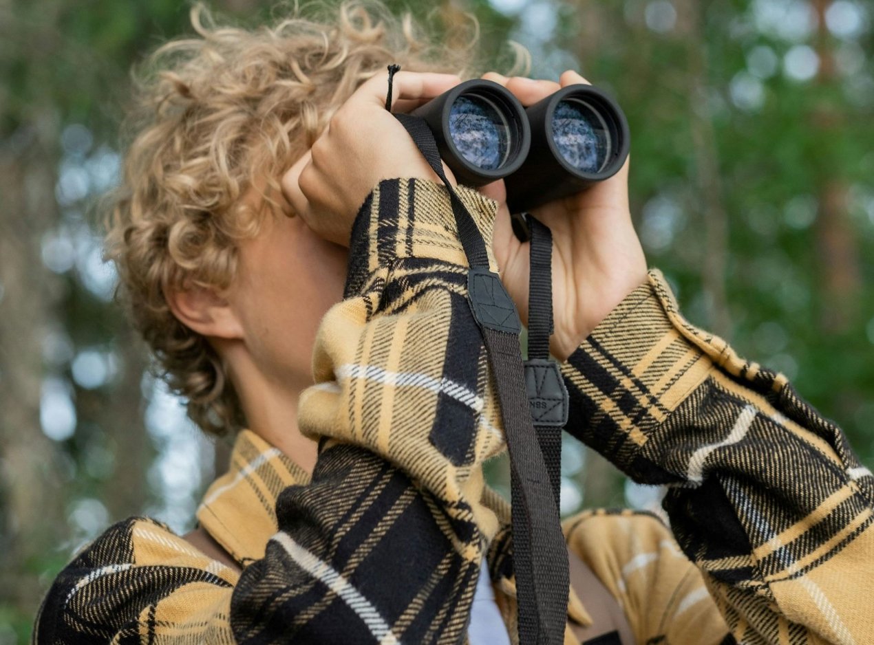 Junge mit Locken schaut durch ein Fernglas und steht im Wald
