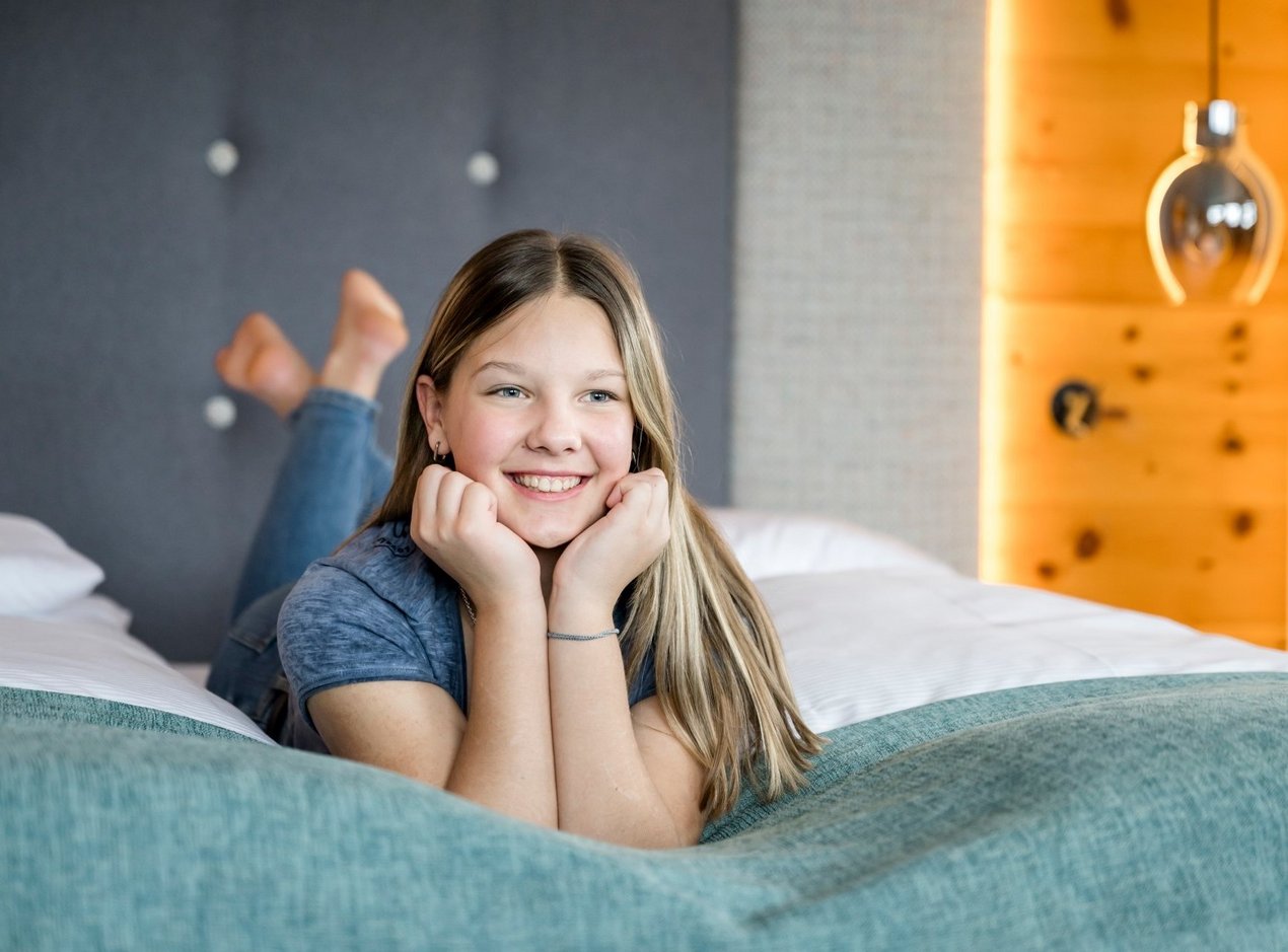 mädche-teenie-im-zimmer Mädchen liegt am Bauch im Bett und strahlt in die Kamera